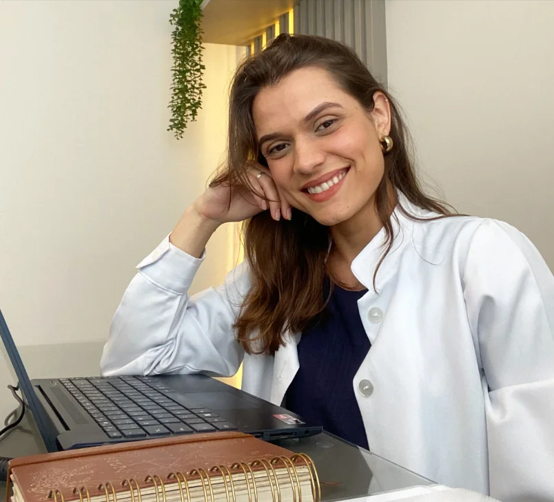 Vitória Avoni de jaleco, sentada à mesa do consultório